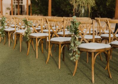 Rows of Wood Cross Back chairs, the chairs have white cushions and are draped with eucalyptus garland