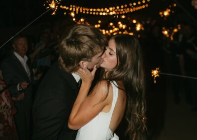 A bride and groom kissing at their reception exit with string lighting and sparklers in the background