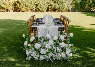 Wood farm table set at an outdoor wedding with wood cross-back chairs and white linen and florals